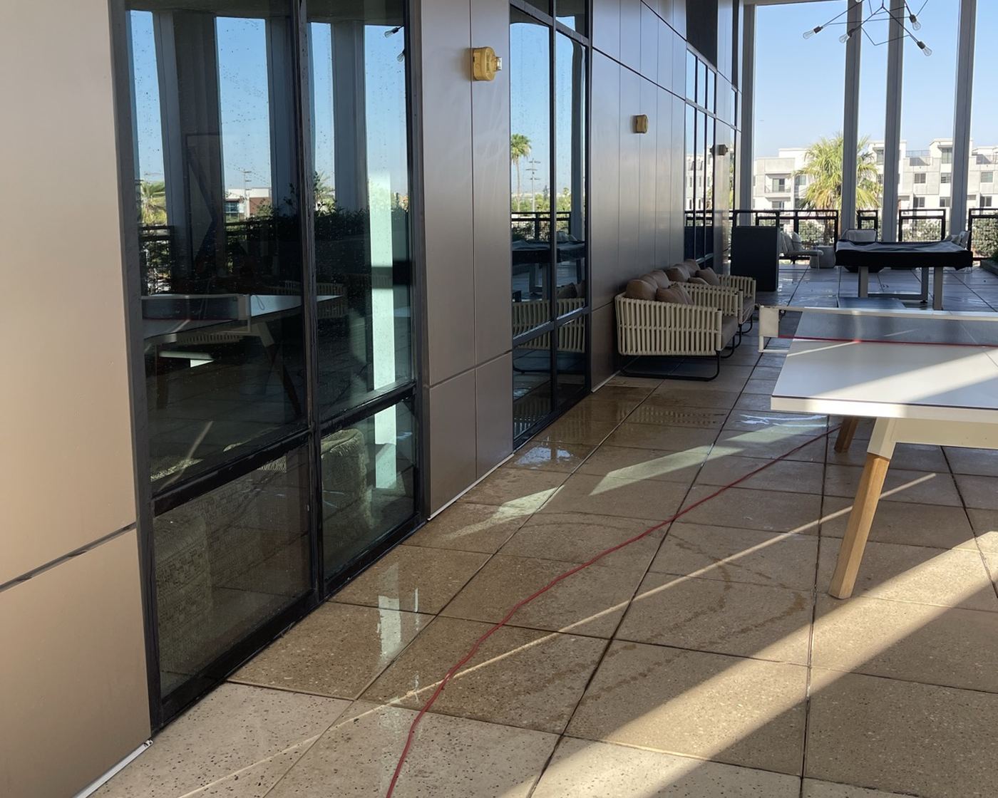 A commercial terrace in Phoenix mid-wash, tile wet and a pressure hose running across it beside a glass curtain wall